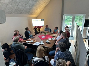 Auf dem Bild ist eine Kleingruppe mit 13 Menschen rund um einen Gruppentisch zu sehen. An der hinteren linken Ecke des Tisches steht ein TV-Bildschirm, auf den die Anwesenden schauen. Der Großteil sitzt mit dem Rücken zur Kamera.  Auf dem Tisch stehen Kaffeetassen und Kuchenteller. Rechts vom Tisch ist eine graue Trennwand zu sehen.&nbsp;&copy;&nbsp;Samtgemeinde Bodenwerder-Polle