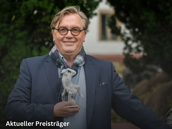 Zu sehen ist Hape Kerkeling, der einen dunkelblauen Anzug mit hellblauem Hemd sowie einen Schal trägt. Rechts in noch ein Teil der Figur des Münchhausen-Brunnens, mit Münchhausen auf seinem halben Pferd zu sehen. Hape Kerkeling hat im Jahr 2025 den Münchhausen-Preis erhalten. Hierfür hat er eine weiße Porzellanfigur von dem Baron auf der Kugel erhalten, welche er hier in der linken Hand hält.&nbsp;&copy;&nbsp;Münchhausenstadt Bodenwerder/Thomas Göbert