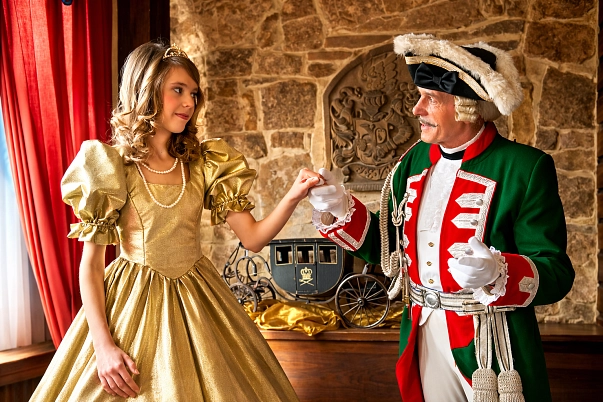 Auf dem Bild sind der Baron von Münchhausen und Aschenputtel zu sehen. Aschenputtel steht links in einem goldenen Kleid und hat eine kleine Krone auf dem Kopf und eine Perlenkette um den Hals. Ihre linke Hand hält der Baron von Münchhausen, der in seiner Uniform mit weißer Hose, grün-roter Jacke und seinem Dreispitz ihre Hand hält. Die beiden schauen sich in die Augen. Hinter Aschenputtel sind ein roter Vorhang und ein Fenster zu sehen. Zwischen den beiden steht im Hintergrund eine dunkle Kommode mit einer kleinen Kutsche vor einer steinernen Wand.&nbsp;&copy;&nbsp;Deutsche Märchenstraße e.V., Fotograf Paavo Blåfield / bearbeitet mit KI