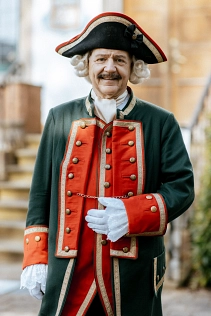 Zu sehen ist der Baron von Münchhausen, der vor dem heutigen Rathaus in Bodenwerder steht. Er trägt seine Uniform mit roter Weste sowie seiner Jacke darüber. Beides hat goldenen Leisten sowie Knöpfe. Die Jacke ist dunkelgrün und rot an den Ärmeln sowie vorne an der offenen Seite. An den Ärmeln schaut ein weißer Spitzenstoff raus und er trägt weiße Handschuhe. Am Hals trägt er ein helles Seidentuch. Auf dem Kopf trägt er einen Dreispitz, der schwarz mit goldener und roter Bordüre ist.  Seine linke Hand hält er vor dem Bauch. Er lächelt freundlich in die Kamera.&nbsp;&copy;&nbsp;SVR/SpiekerFotografie, CC BY, Solling-Vogler-Region im Weserbergland