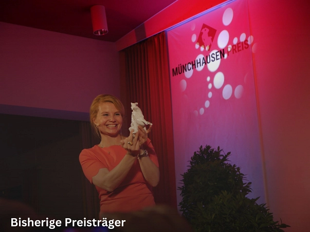 Zu sehen ist Annette Frier in einem orangen Kleid. Sie hält den Münchhausen-Preis, eine weiße Porzellan-Figur mit dem Baron von Münchhausen bei seinem berühmten Kugelflug, stolz in der Hand und zeigt ihr nach oben. Dabei lächelt sie freudig. Im Hintergrund ist ein Banner mit dem Schriftzug Münchhausen-Preis zu sehen. Dieser erscheint im lila Licht.&nbsp;&copy;&nbsp;Münchhausenstadt Bodenwerder