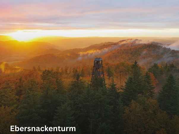 Ebersnackenturm.jpg Zu sehen ist eine Luftaufnahme des Ebersnackenturmes. Eines Turm aus Holz gebaut, der über die Baumkronen des Voglers ragt. Rund um den Turm stehen Bäume. Im Hintergrund legt sich der Nebel über das Tal. Links ist die gerade aufgehende Sonne zu sehen. © SVR/M. Sprödefeld