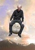 Emil Steinberger sitzt lächelnd auf der Kanonenkugel und „fliegt“ über durch den bewölkten Himmel über den Bergen. Er trägt ein schwarzes langes Oberteil, eine schwarze lange Hose und schwarze Schuhe.&nbsp;&copy;&nbsp;Tom Freitag