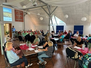 Auf dem Bild sind einige Menschen sitzend an verschiedenen Gruppentischen zu sehen. Sie sitzen in einem großen Raum mit hoher Decke. Unter der weißen Decke hängen einige Lampions und Tücher zur Deko. Die Menschen schauen in Richtung der Vorderseite des Raumes, welche hier auf der linken Seite zu sehen ist. Dort wird ein Bild an eine Leinwand geworfen und es steht ein junger Mann dort und redet zu den Menschen.&nbsp;&copy;&nbsp;Samtgemeinde Bodenwerder-Polle