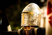 Auf dem Bild steht ein goldener Ritterhelm im Fokus. Der Hintergrund verschwimmt größtenteils, links ist grüne Natur zu sehen und im rechten Teil hinter dem Helm ein Fachwerkgebäude sowie eine rote Fahne. Unter dem Helm liegt ein Kettenhemd sowie weitere Teile einer Ritterrüstung.&nbsp;&copy;&nbsp;Samtgemeinde Bodenwerder-Polle - Die Photo-WG