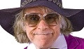 Auf dem Bild ist eine Nahaufnahme von Helge Schneiders Kopf zu sehen. Dieser verzieht sein Gesicht zu einem leichten Grinsen. Sein Grau-blondes Harr ist beinahe schulterlang und er trägt einen schmalen Oberlippenbart. Helge Schneider trägt eine eckige Sonnenbrille mit einem durchsichtigen Gestell. Die Bügel sind gold. Durch die Gläser sind seine Augen zu sehen. Auf dem Kopf trägt er einen dunkelgrauen Filzhut mit einem Kranz aus kleinen Filzblättchen mit goldenen Verzierungen und einer großen Krempe. Zudem ist der Kragen seines weißen Hemdes mit fliederfarbenen Streifen zu sehen. Dazu trägt er eine pinkfarbene Cordjacke.&nbsp;&copy;&nbsp;Helge Schneider