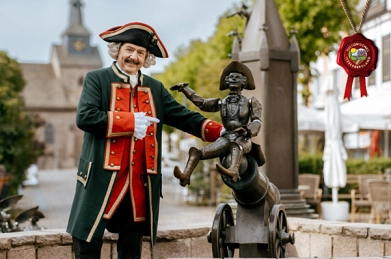 Auf dem Bild ist der Baron von Münchhausen in seiner Uniform am Münchhausen-Brunnen in der Fußgängerzone in Bodenwerder zu sehen. Er trägt eine dunkle Hose und eine dunkelgrüne lange Jacke mit roten sowie goldenen Details sowie goldenen Knöpfen vorne. Auf dem Kopf trägt wer einen Dreispitz mit ebenfalls roten und goldenen Verzierungen an den Kanten. Am Hals trägt er einen weißen Einsatz. An den Ärmeln schaut weiße Spitze hervor. Außerdem trägt er weiße Handschuhe. Er zeigt auf die Figur des Münchhausen-Brunnes, die den Flug auf der Kanonenkugel darstellt. Im Hintergrund ist noch die Figur zu sehen, die die Geschichte mit dem Pferd am Kirchturm erzählt. Hinter dem Brunnen sind unscharf ein Teil der Fußgängerzone sowie die Klosterkirche St. Nicolai zu sehen.&nbsp;&copy;&nbsp;CC BY SVR_SpiekerFotografie/Siegel: Weserbergland Tourismus e.V.