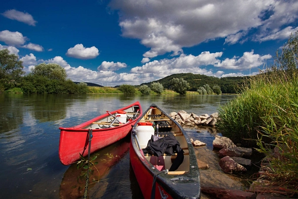 Im Vordergrund liegen ein rotes und ein schwarzes Kanu am Weserufer. Im Hintergrund sind die Weser sowie Bäume und Gräser zu sehen.&nbsp;&copy;&nbsp;Samtgemeinde Bodenwerder-Polle - Rolf Sander