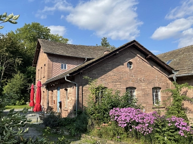 Auf dem Bild ist die KulturMühle sowie das dazugehörige MühlenCafé in Buchhagen zu sehen. Die historische Sandsteinmühle wurde 1867 erbaut, seit 1908 ist der Schleifbetrieb eingestellt. Links vor dem Gebäude stehen zwei zusammengeklappte rote Sonnenschirme sowie ein paar Stühle mit Tisch. Im Vordergrund sind einige grüne Büsche und Bäume zu sehen sowie auf der rechten Seite lila blühende Blumen.&nbsp;&copy;&nbsp;Samtgemeinde Bodenwerder-Polle - Simone Kelly