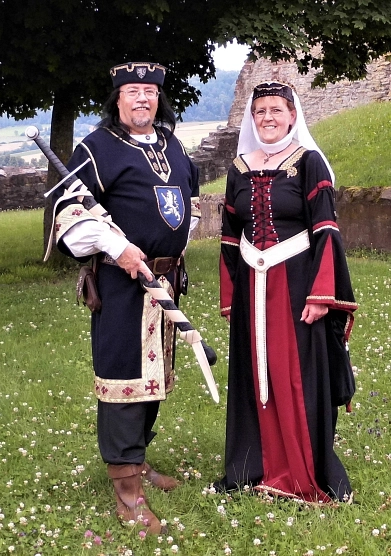 Auf dem Bild sind die Darsteller des Graf Otto von Everstein und seiner Gemahlin Lutgardis zu sehen. Diese stehen auf einer Rasenfläche, im Hintergrund sind ein Teil der Burgruine Polle sowie ein Baum zu sehen. Graf Otto von Everstein steht auf der linken Seite und trägt ein schwarzes Gewand mit einer hellen Bordüre an den Ärmeln und unten. Vorne drauf ist das blau-weiße Wappen der Eversteiner zu sehen. Dazu trägt er eine Schwarze Hose und braune Stiefel. Zudem einen braunen Gürtel mit einer kleinen Tasche. Unter dem Gewand trägt er ein weißes Hemd und einen passenden schwarzen Hut, ebenfalls mit Wappen, auf dem Kopf. In der Armbeuge hält er ein Schwert mit der Spitze nach unten. Graf Otto von Everstein hat schwarze lange Haare und eine Brille. Er lächelt freundlich in die Kamera. Rechts von ihm steht seine Gemahlin Lutgardis. Diese trägt ein schwarz-rotes langes Kleid mit einem weißen Gürtel um die Hüfte. Vorne hat das Kleid eine Schnürung und goldene Verzierungen am Ausschnitt und an den Ärmeln. Sie trägt eine schwarze Kappe auf dem Kopf, mit einem weißen Schleier. Auch sie lächelt ebenfalls freundlich in die Kamera.&nbsp;&copy;&nbsp;Samtgemeinde Bodenwerder-Polle - Andreas Busche