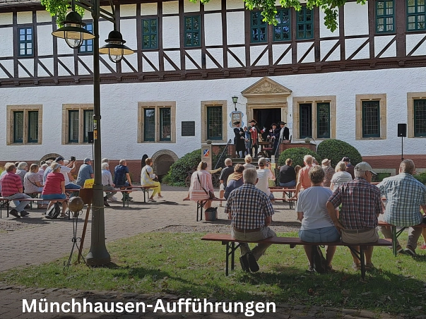 Im Vordergrund sind Menschen zu sehen, die mit dem Rücken zur Kamera auf Holzbänken sitzen und Richtung Rathaus schauen. Das weiße Fachwerkgebäude ist im Hintergrund zu sehen. Dort auf dem Plateau an der Tür stehen ein Holztisch sowie Holzstühle. Hier oben ist die Münchhausen-Spielgruppe mit dem Darsteller des Baron von Münchhausen in seiner Uniform zu sehen. Dies zeigt die Aufführung des Münchhausen-Spiels.&nbsp;&copy;&nbsp;Münchhausenstadt Bodenwerder