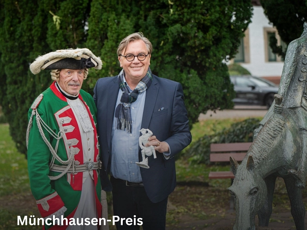 Zu sehen ist auf der linken Seite der Darsteller des Baron von Münchhausen, der seine grün-rote Uniform sowie weißer Hose, Weste und einen Dreispitz trägt. Direkt daneben steht Hape Kerkeling, der einen dunkelblauen Anzug mit hellblauem Hemd sowie einen Schal trägt. Rechts in noch ein Teil der Figur des Münchhausen-Brunnens, mit Münchhausen auf seinem halben Pferd zu sehen. Hape Kerkeling hat im Jahr 2025 den Münchhausen-Preis erhalten. Hierfür hat er eine weiße Porzellanfigur von dem Baron auf der Kugel erhalten, welche er hier in der linken Hand hält.&nbsp;&copy;&nbsp;Münchhausenstadt Bodenwerder/Thomas Göbert