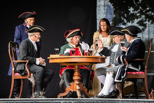 Münchhausenspielgruppe Das Münchhausenspiel zeigt den Baron von Münchhausen im Kreise seiner Freunde. Sie sitzen am runden, braunen Holztisch und lauschen angeregt seinen Erzählungen bei einem Gläschen Punsch.
Bewirtet von seiner Haushälterin, Frau Nolte berichtet er von der Zeit als Offizier und Rittmeister in Russland. Seine unverwechselbare Fabulierkunst stellt er gerne unter Beweis.
In der Mitte des Tisches sitzt der Baron von Münchhausen und erzählt seine Geschichten. Er trägt einen grünen Anzug mit breitem rotem Revers, welches mit goldenen Knöpfen versehen ist. An den Vorderkanten von Rock und Weste laufen goldene Stickereien entlang und umrahmen die Taschen. Dazu trägt er schwarze Lederstiefel und einen schwarzen Dreispitz, welcher goldene und rote Verzierungen hat.
Unter dem Anzug ragt der weiße Spitzenbesatz der Ärmel hervor. Als Einziger trägt der Baron weiße Handschuhe.
Wie zu der Zeit üblich trägt die Haushälterin, Frau Nolte, einen Reifrock. Darüber trägt sie einen Rock und ein einteiliges vorne offenes Kleid mit hellem Brusteinsatz.
Die Ärmel sind ellenbogenlang und enden in flügelartigen Aufschlägen. Das Kleid zieren hellbeige Rüschen und Stickereien. Um die Hüfte gebunden trägt die Haushälterin eine weiße Schürze. In der rechten Hand hält sie einen gefüllten Tonkrug mit rotem Punsch.
Um den Tisch versammelt sitzt rechts vom Baron der Pastor in einem schwarzen Anzug mit Weste und Kniehose mit schwarzen Kniestrümpfen An den Handgelenken, sowie auf der Brust ist der Spitzenbesatz des Hemdes zu sehen. Dazu trägt er eine weiße Halsbinde und schwarze Lederschuhe mit goldener Schnalle. Auf dem Kopf trägt er einen Dreispitz mit goldenen Akzenten. Hinter dem Pastor steht Herr Rösemeier in einem blauen Anzug mit schwarzem Dreispitz und roten Akzenten.
Links neben dem Baron sitzt der Amtmann, dieser trägt einen grauen Anzug mit grauer Weste und einer weißen Halsbinde. Auch hier ist der Spitzenbesatz der Ärmel zu sehen. Der Amtmann trägt weiße Kniestrümpfe und seinen schwarzen Dreispitz zieren silberne Akzente. Links neben dem Amtmann sitz Dr. Krusins in einem schwarzen Anzug mit schwarzer Weste und grauer Kniehose. Dazu trägt er weiße Kniestrümpfe und schwarze Lederschuhe mit silberner Schnalle. Er führt sein Glas Punsch zum Mund. © Joachim Merker