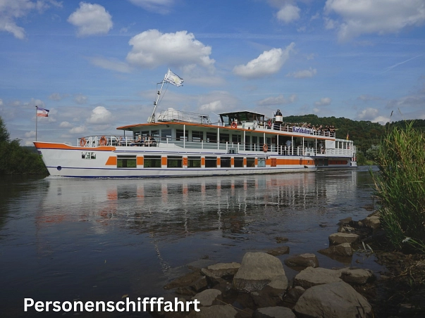 Das Bild wurde vom Weserufer aus aufgenommen. Direkt im Mittelpunkt ist ein Personenschiff, welches weiß mit einigen orangen Akzenten ist. Das Schiff trägt den Namen "Karlshafen". Oben an Deck stehen einige Mitfahrer und schauen in die Landschaft, auch im vorderen Bereich des Schiffes sitzen einige Mitfahrer. Es sind zwei Flaggen im vorderen Bereich des Schiffes gehisst. Eine davon ist von der Flotte Weser GmbH, dem Betreiber der Personenschifffahrt auf der Weser. Links und rechte im Bild sind Grünpflanzen zu sehen, die direkt am Weserufer wachsen.&nbsp;&copy;&nbsp;Samtgemeinde Bodenwerder-Polle/Rolf Sander