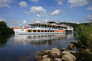 Das Bild wurde vom Weserufer aus aufgenommen. Direkt im Mittelpunkt ist ein Personenschiff, welches weiß mit einigen orangen Akzenten ist. Das Schiff trägt den Namen "Karlshafen". Oben an Deck stehen einige Mitfahrer und schauen in die Landschaft, auch im vorderen Bereich des Schiffes sitzen einige Mitfahrer. Es sind zwei Flaggen im vorderen Bereich des Schiffes gehisst. Eine davon ist von der Flotte Weser GmbH, dem Betreiber der Personenschifffahrt auf der Weser. Links und rechte im Bild sind Grünpflanzen zu sehen, die direkt am Weserufer wachsen.&nbsp;&copy;&nbsp;Samtgemeinde Bodenwerder-Polle - Rolf Sander