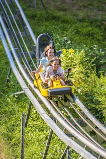 Auf dem Bild ist ein Ausschnitt der Schienen der Rodelbahn zu sehen, mit grünem grün bewachsenem Hintergrund. Links unten ist ein Teil eines Drahtzaunes zu sehen. Auf der Bahn rodeln gerade zwei Mädchen in einem gelben Bob die Strecke runter. Das vordere Mädchen im weißen T-Shirt hält sich an der entsprechenden Halterung fest und hat ein breites Grinsen auf dem Gesicht. Das hintere Mädchen trägt ein hellblaues T-Shirt und hält sich an den seitlichen Bremsen fest. Auch sie hat ein Lächeln im Gesicht.&nbsp;&copy;&nbsp;Samtgemeinde Bodenwerder-Polle - Carolin Schwiete/Thomas Göbert