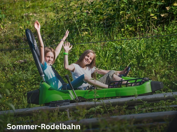 Sommer-Rodelbahn.jpg Zu sehen ist ein grüner Bob der Rodelbahn auf den Schienen. Hierin sitzen zwei Mädchen mit hellen T-Shirts, die lachend in die Kamera winken. © Sommer-Rodelbahn Bodenwerder/C. Schwiete