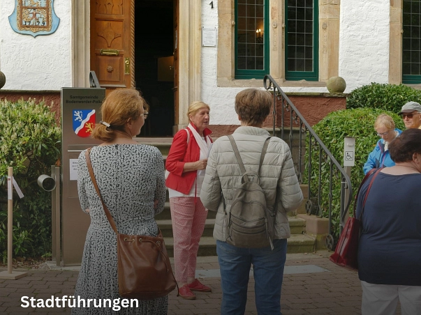 Stadtführungen.jpg Zu sehen ist eine Stadtführerin in roter Jacke und hellroter Hose sowie weißem T-Shirt. Diese steht mit dem Gesicht zu Kamera und steht vor dem Geburtshaus des Baron von Münchhausen, dem heutigen Rathaus. Hinter ihr ist die Steintreppe sowie eine offene Holztür. Mit dem Rücken zu r Kamera stehen einige Teilnehmende der Stadtführung, die in die Richtung der Stadtführerin schauen. © Samtgemeinde Bodenwerder-Polle/Rolf Sander