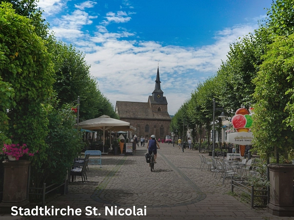Stadtkirche St. Nicolai.jpg Zu sehen ist der Blick von der Fußgängerzone auf die Stadtkirche. Rechts und links umranden dicht bewachsene grüne Bäume die Fußgängerzone. Vereinzelt sind Sonnenschirm oder Beschilderungen der ansässigen Gastronomie zu sehen. Zudem einige Menschen zu Fuß oder mit dem Rad. © Samtgemeinde Bodenwerder-Polle/Rolf Sander