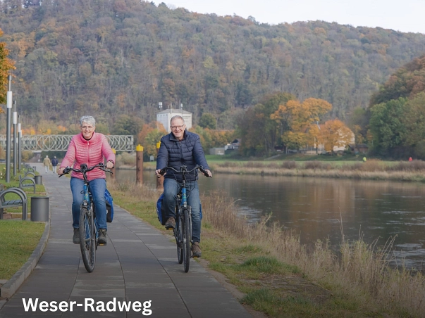 Weser-Radweg.jpg Auf dem Bild ist ein Teil der Weserpromenade in der Münchhausenstadt Bodenwerder zu sehen. Hier fahren auf dem unteren Weg direkt an der Weser zwei Radfahrer. Links eine Frau in einer blauen Jeans uns einer pinkfarbenen Steppjacke, rechts ein Mann in einer Jeans und einer dunkelblauen Steppjacke. Beide haben blaue Satteltaschen an ihren Rädern und fahren auf die Kamera zu. Außerdem schauen beide in die Kamera und lächeln. Die Weser befindet sich rechts von den Radfahrern, links von ihnen Laternen und Bänke entlang des Weges. Im Hintergrund ist die andere Weserseite, eine Eisenbahnbrücke und der Blick auf den bewaldeten Eckberg zu sehen. Das Bild wurde im Herbst aufgenommen. © CC BY-SA: SVR/KoernerPaeslack