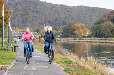 Auf dem Bild ist ein Teil der Weserpromenade in der Münchhausenstadt Bodenwerder zu sehen. Hier fahren auf dem unteren Weg direkt an der Weser zwei Radfahrer. Links eine Frau in einer blauen Jeans uns einer pinkfarbenen Steppjacke, rechts ein Mann in einer Jeans und einer dunkelblauen Steppjacke. Beide haben blaue Satteltaschen an ihren Rädern und fahren auf die Kamera zu. Außerdem schauen beide in die Kamera und lächeln. Die Weser befindet sich rechts von den Radfahrern, links von ihnen Laternen und Bänke entlang des Weges. Im Hintergrund ist die andere Weserseite, eine Eisenbahnbrücke und der Blick auf den bewaldeten Eckberg zu sehen. Das Bild wurde im Herbst aufgenommen.&nbsp;&copy;&nbsp;CC BY-SA: SVR/KoernerPaeslack